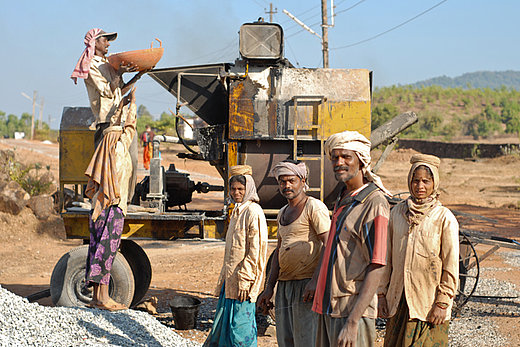 Frauen und Maenner transportieren die Steine zu einer Maschine, die diese zerkleinert.  Kein Foerderband erleichtert die Arbeit, die Behaeltnisse muessen von Hand zu Hand gereicht werden.